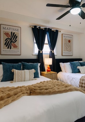 Ein Hotelzimmer mit zwei Betten, dunklen Vorhängen, einem Deckenventilator und zwei an der Wand befestigten Kunstwerken.
