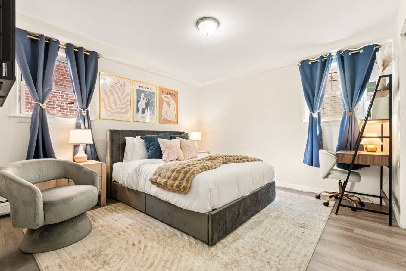 Un dormitorio moderno con una cama grande, un escritorio y una silla. Hay cortinas en las ventanas y una obra de arte en la pared.