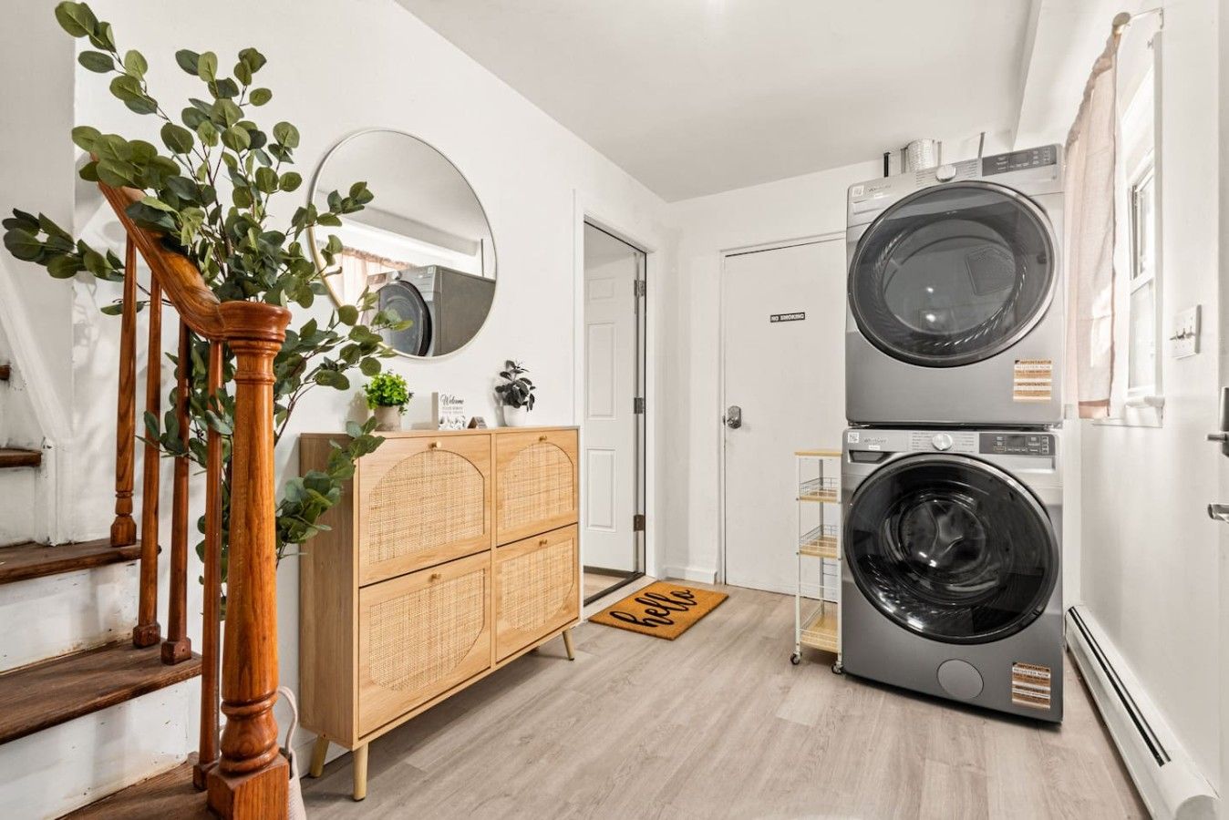 A laundry room with two stacked washing machines, a wooden cabinet, a mirror, and a staircase with a plant.