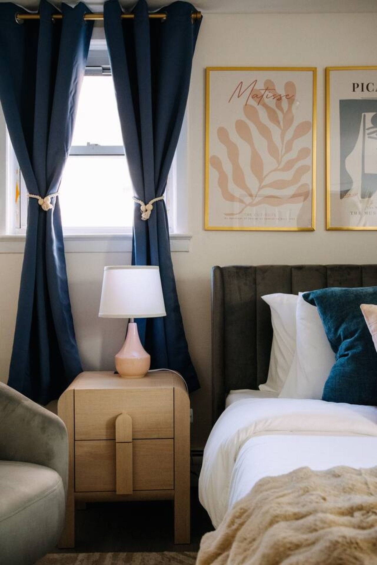 A hotel room with a bed, bedside table, lamp, curtains, and two framed artworks on the wall.