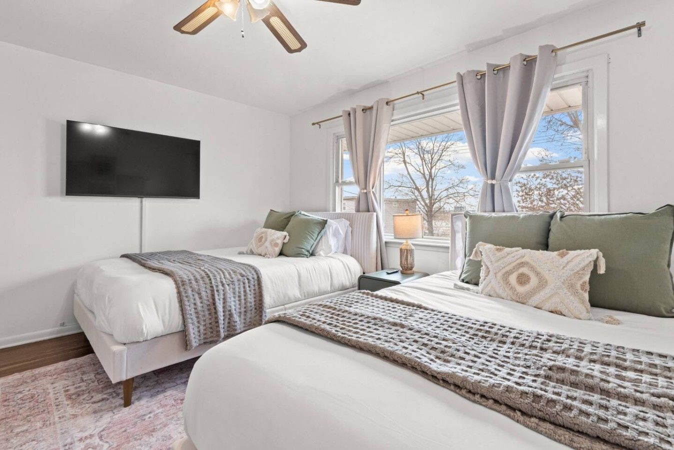 Un dormitorio con una cama grande, una cama más pequeña, un televisor de pantalla plana en la pared y una ventana con cortinas.