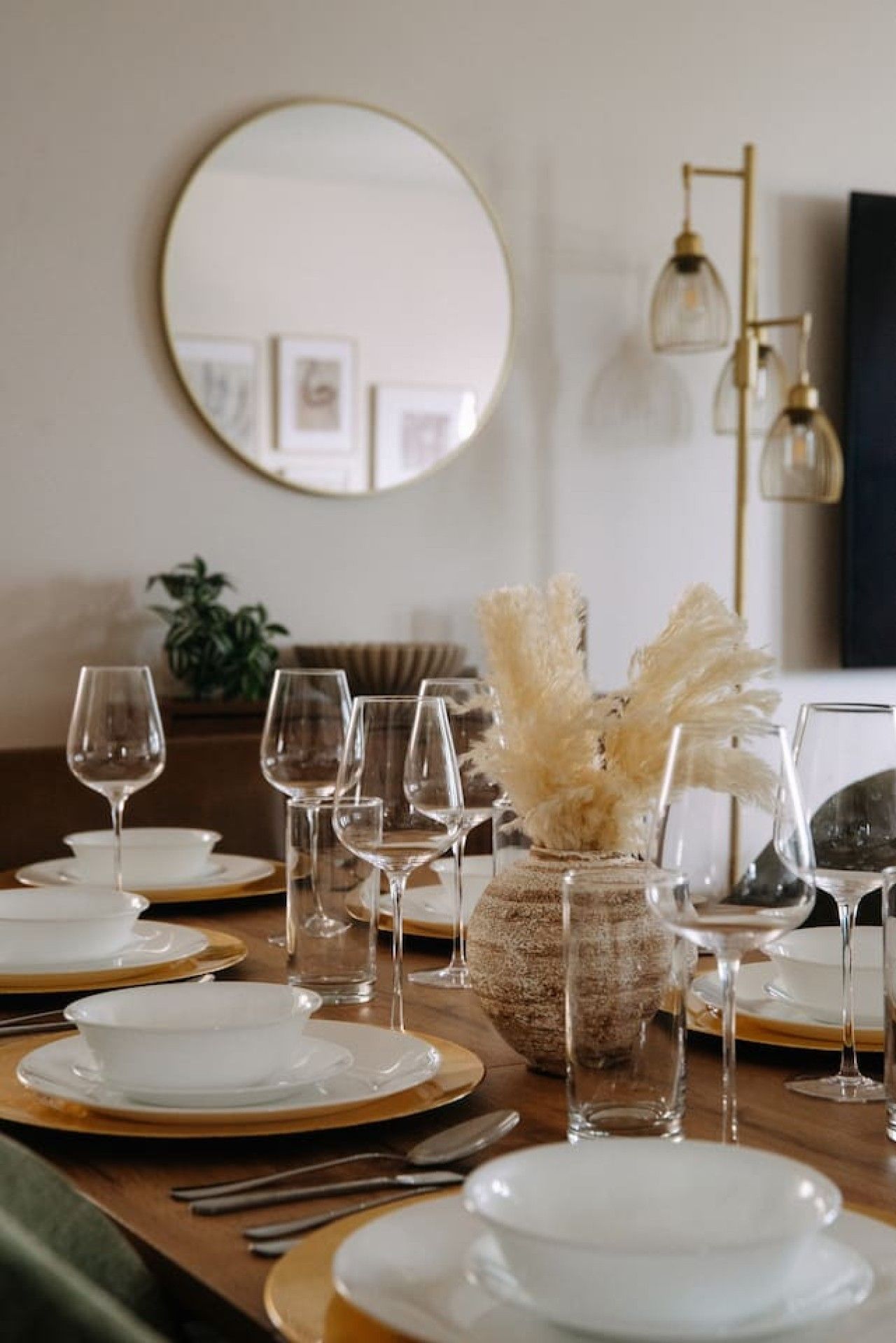 A dining table set with white plates and gold accents, a vase with dried flowers, and a gold-framed mirror on the wall.