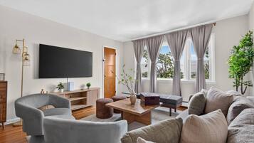 A modern living room with a flat-screen TV, grey sofas, a wooden coffee table, and a large window with curtains.
