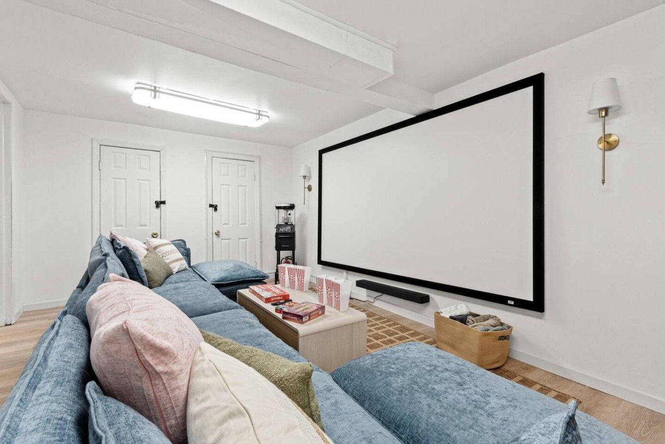 A modern living room with a large flat-screen TV mounted on the wall, a sofa with multiple cushions, a coffee table with snacks, and a projector stand.