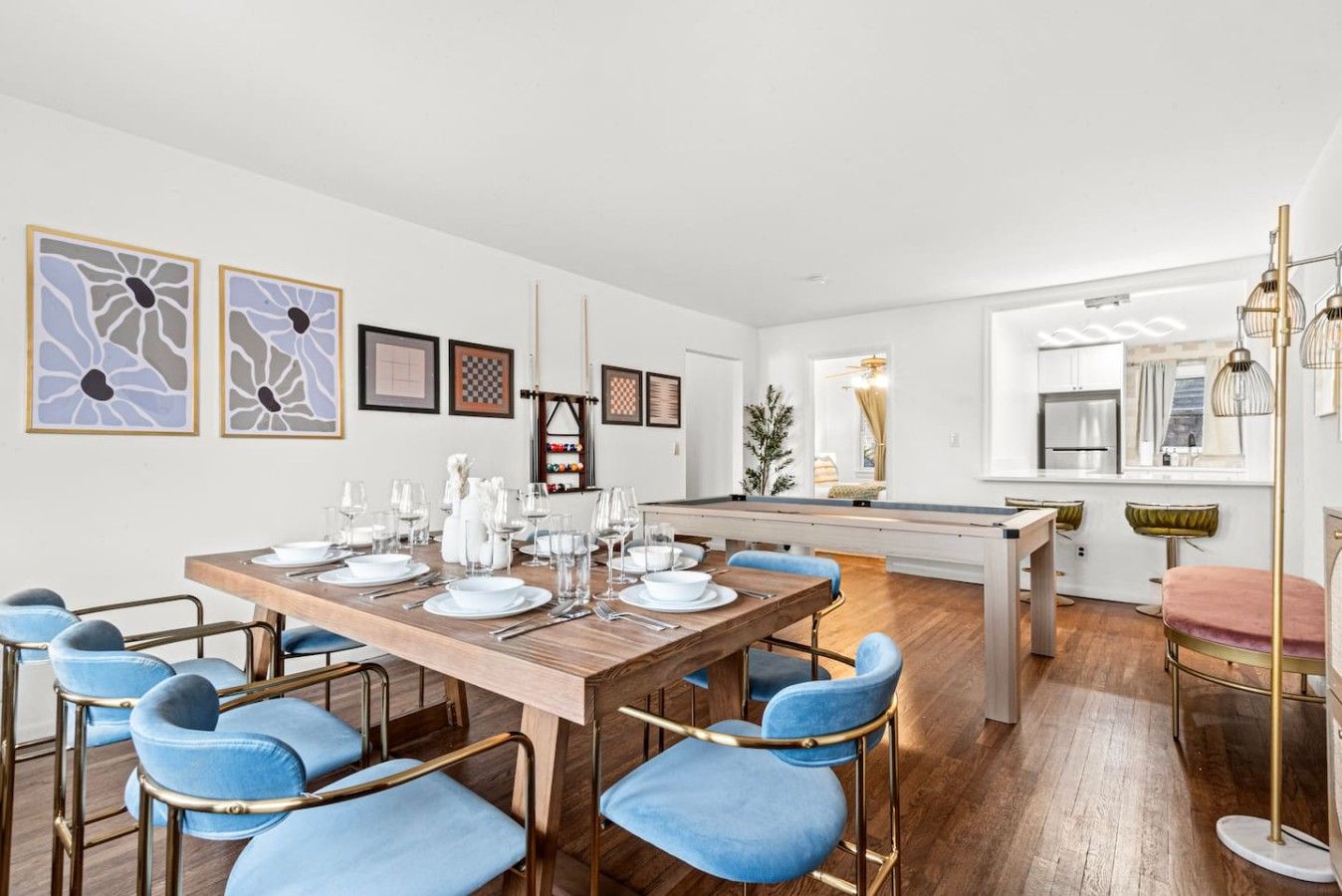 A modern dining area with a wooden table set for six, blue upholstered chairs, and a kitchen visible in the background.