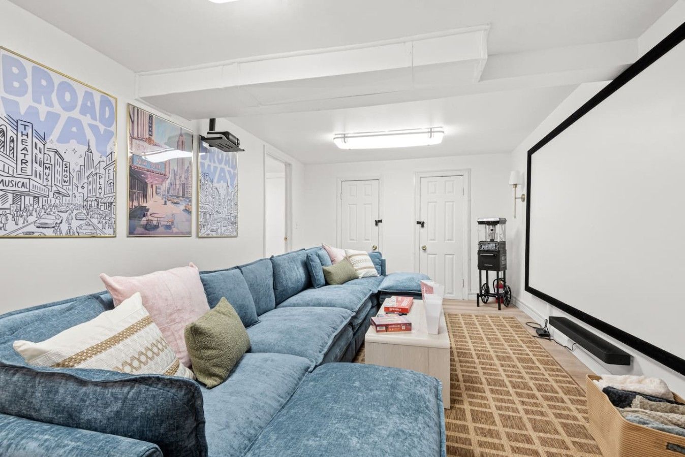 A living room with a sectional sofa, a coffee table, a projector screen, and framed posters on the wall.