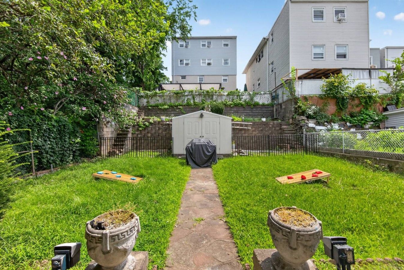Un jardin avec un chemin pavé menant à un abri de jardin, le tout entouré d’une clôture et de verdure.