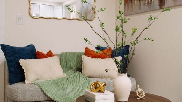 A living room with a grey sofa, multiple cushions, a round coffee table with decorative items, a painting on the wall, and a ceiling fan. - Big City Retreat with Parking (North Bergen)