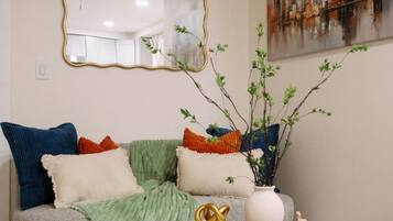 A living room with a grey sofa, multiple cushions, a round coffee table with decorative items, a painting on the wall, and a ceiling fan.