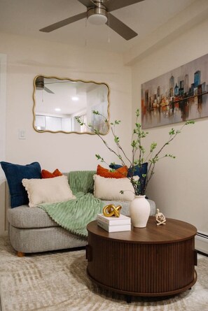 Sala de estar com sofá cinza, várias almofadas, mesa de centro redonda com objetos decorativos, um quadro na parede e um ventilador de teto.