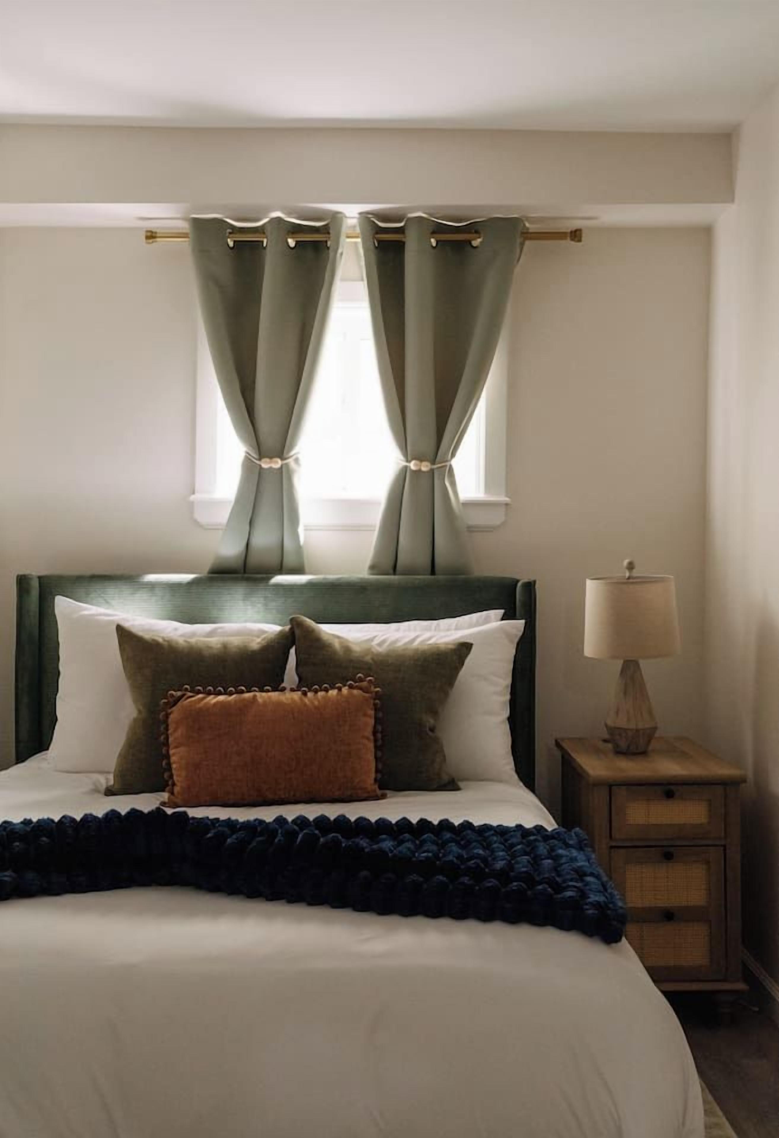 Una cama bien tendida con cabecera, lámpara de noche y ventana con cortinas.