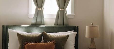 Una cama bien tendida con cabecera, lámpara de noche y ventana con cortinas.