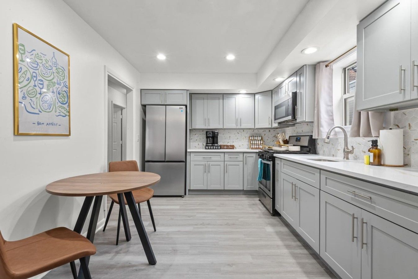 Una cocina moderna con gabinetes grises, un refrigerador de acero inoxidable, un microondas y una estufa. Hay un comedor con una mesa redonda y dos sillas.