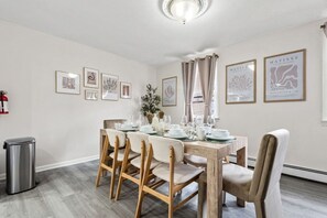 Uma área de refeições com uma mesa de madeira posta para quatro pessoas, rodeada por cadeiras, com quadros nas paredes e um extintor de incêndio à esquerda.