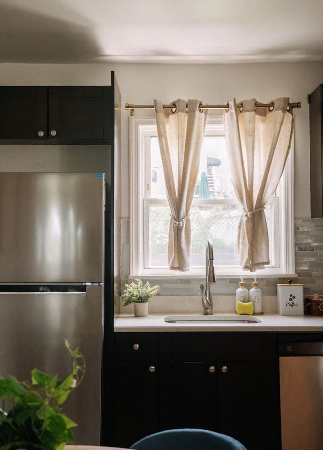 A kitchen with a stainless steel refrigerator, a sink with a modern faucet, and a window with beige curtains.