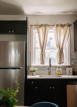 Eine Küche mit einem Edelstahlkühlschrank, einem Spülbecken mit einem modernen Wasserhahn und einem Fenster mit beigen Vorhängen.