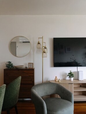 Ein Wohnzimmer mit einem runden Spiegel, einem Wand-Fernseher, einer Stehlampe, einem Holzschrank und zwei Sesseln.