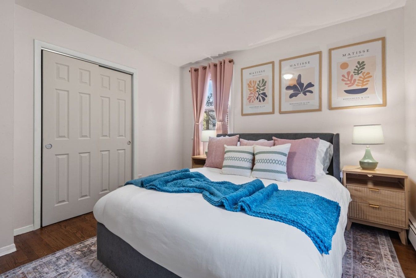 A bedroom with a bed, a nightstand, a lamp, a door, and three framed artworks on the wall.