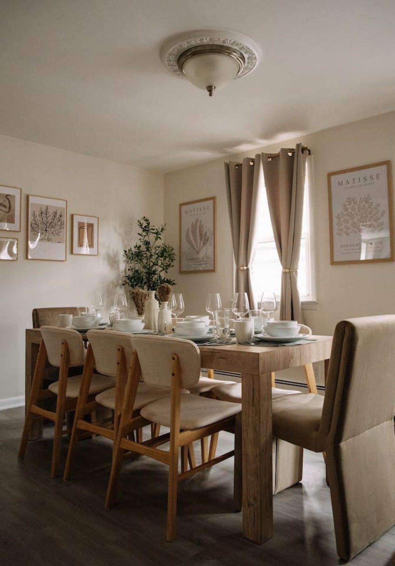 A dining room with a wooden table set for four, surrounded by chairs, with framed artwork on the walls and a window with curtains.