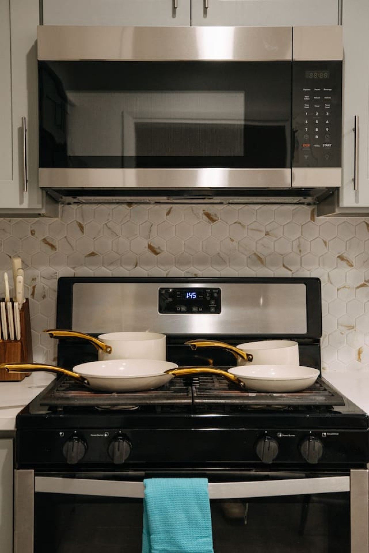 Una cocina con un microondas de acero inoxidable, una estufa con dos ollas y un reloj digital.