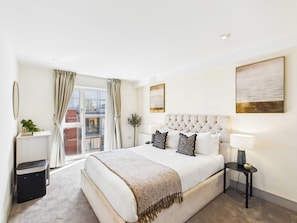 A hotel room with a large bed, a bedside table with a lamp, a window with curtains, and artwork on the walls. - Westminster Duplex 2BR Apartment with Balcony (Greater London)