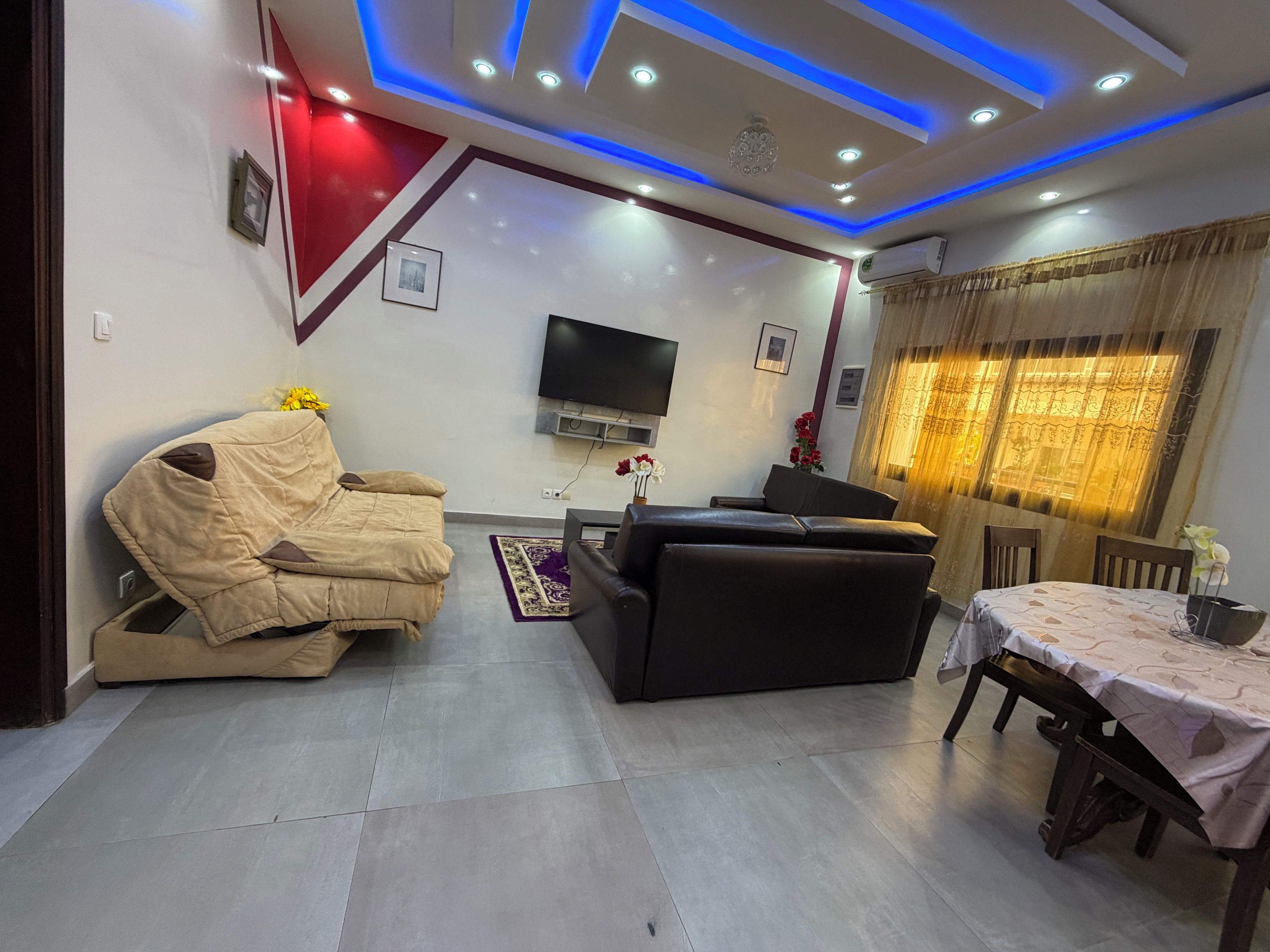 A modern living room with a sofa, a television mounted on the wall, and a dining area with a table and chairs.