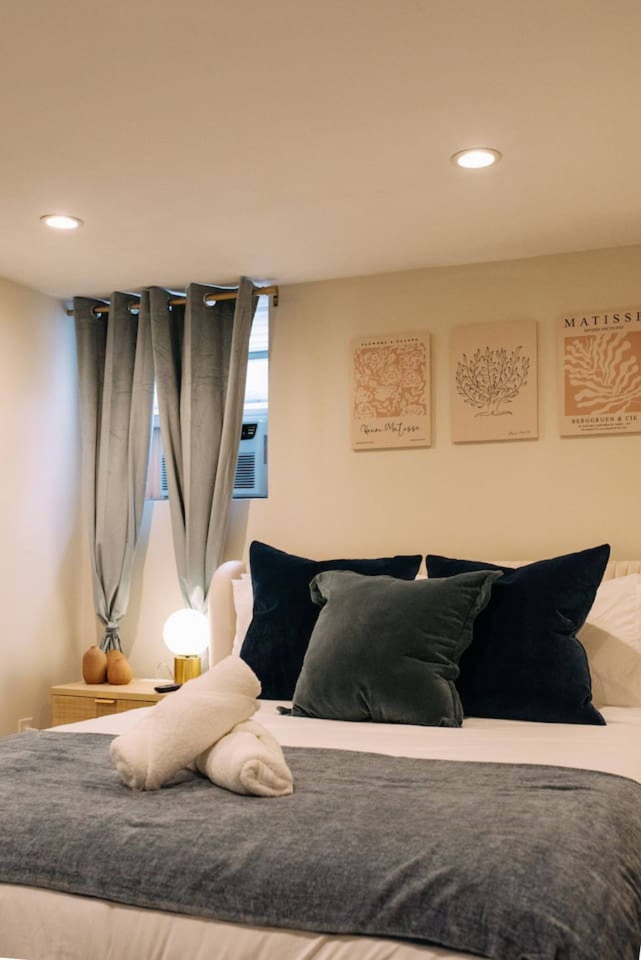 Una cama bien hecha con almohadas y una colcha, una mesita de noche con lámpara y una ventana con cortinas.