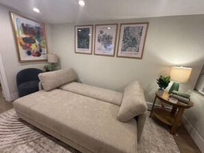 Una habitación con un sofá, una silla, una mesita de noche con una lámpara y cuadros en la pared.
