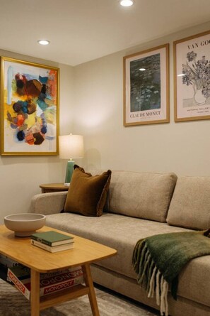 A living room with a beige sofa, a small table with books, a bowl, and a lamp. There are framed artworks on the wall. - Chic Luxe mins NYC & MetLife W/ Private Backyard (Hoboken)