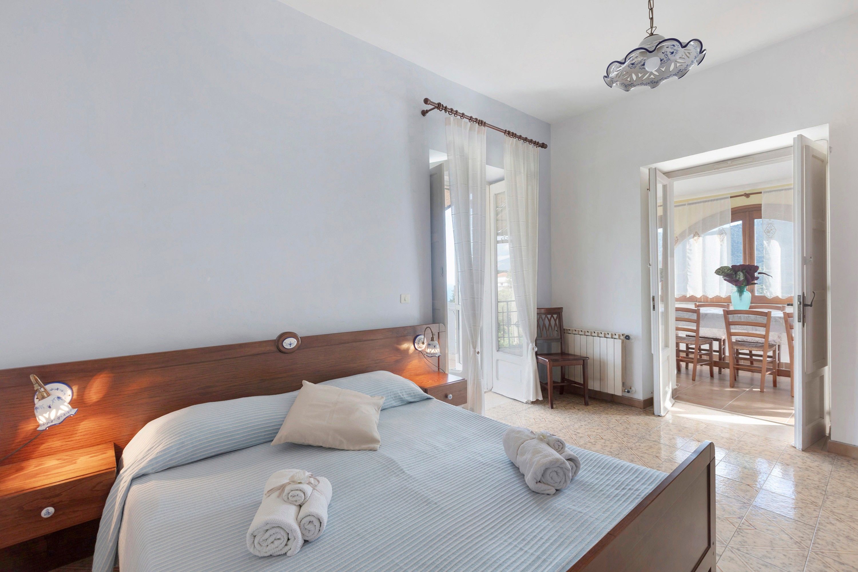 A hotel room with a bed, bedside tables, a hanging light fixture, a view of the dining area, and a door leading outside.