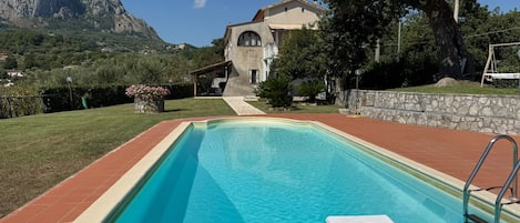 A swimming pool with a stone wall and a metal ladder, surrounded by a grassy area and a building with a mountain in the background.