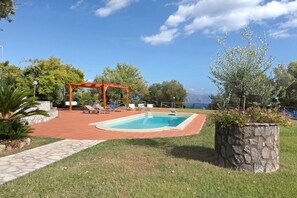 Ein Swimmingpool mit einer roten Pergola und Liegestühlen, umgeben von Gras und Bäumen.