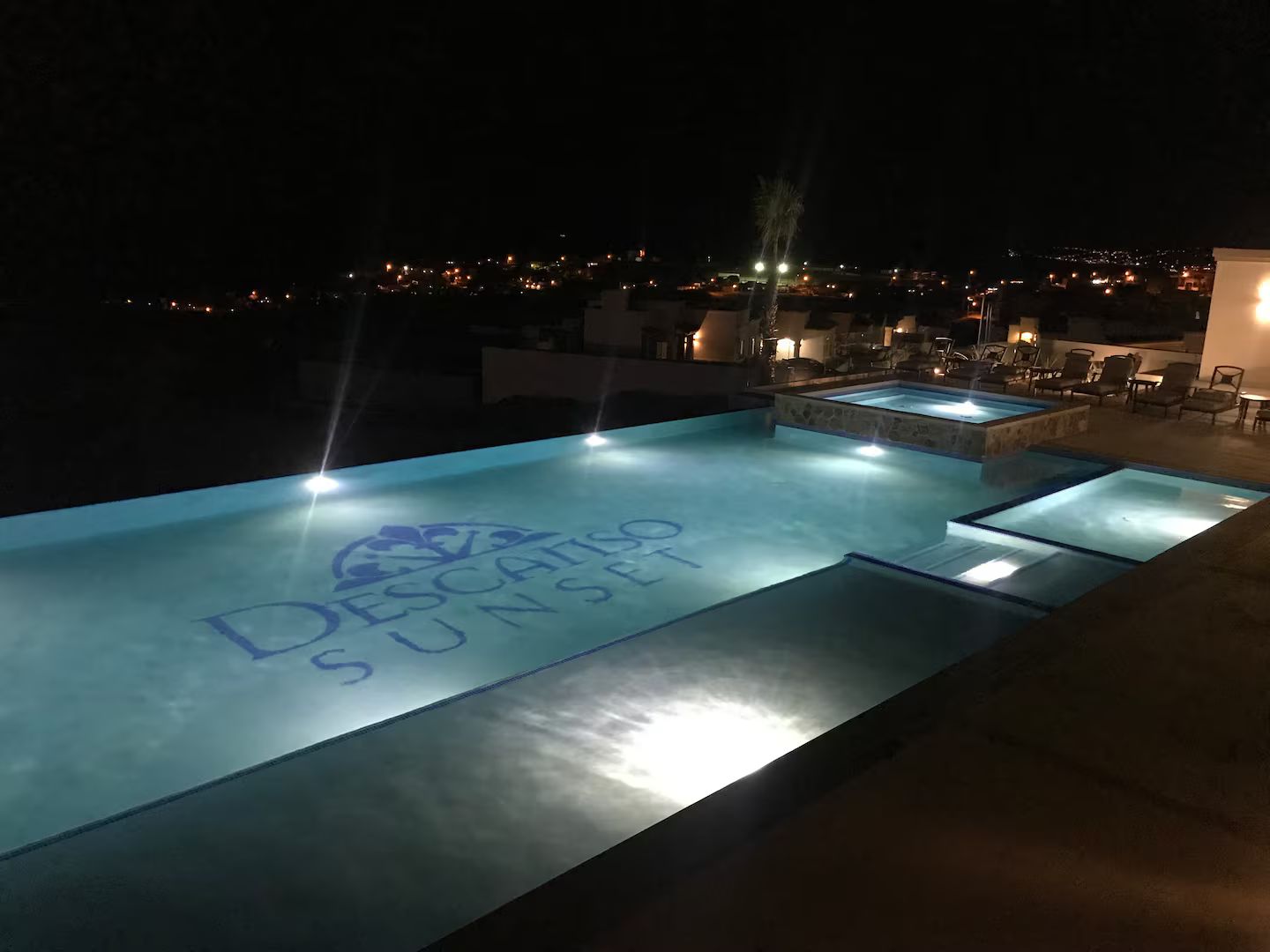 An outdoor infinity pool with 'DESCARTES SUNSET' written on the water, surrounded by lounge chairs and a cityscape view at night.