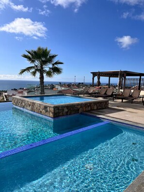 Ein klares, blaues Schwimmbecken mit Steinumrandung, einer Palme, Liegestühlen und Blick aufs Meer.