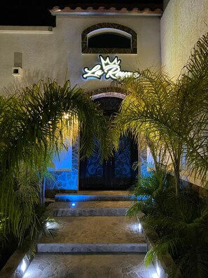 Un ingresso con insegna al neon, una scala in pietra e piante tropicali.