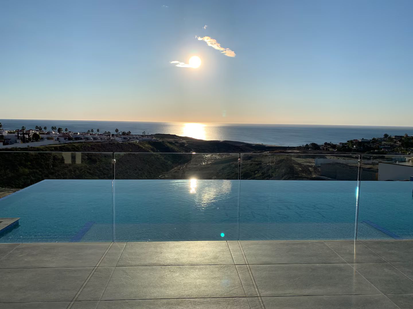 A clear view of a swimming pool with a glass barrier, overlooking a coastal landscape with buildings and the ocean.