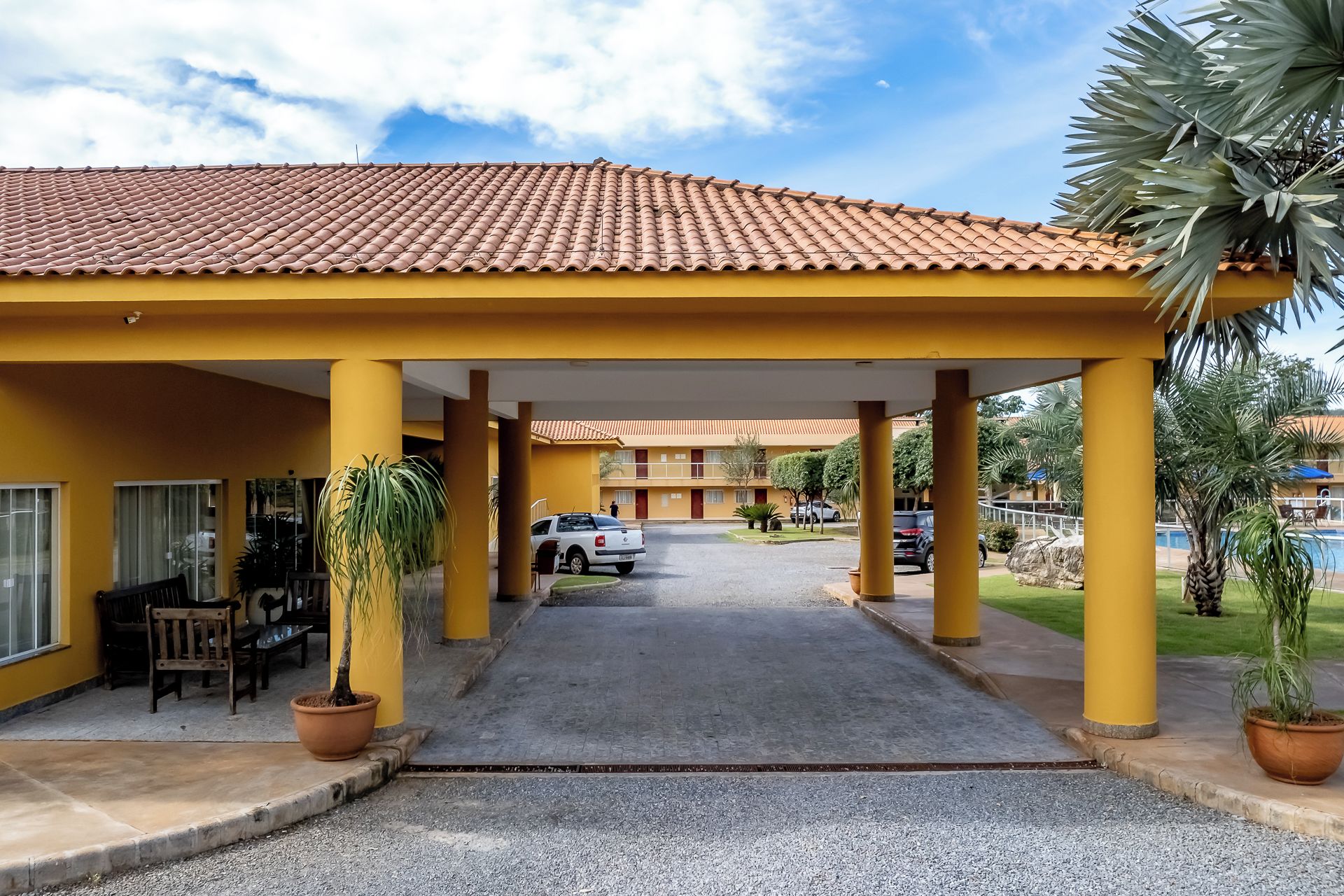 A hotel with a covered entrance, a parking area, and a pool in the background.