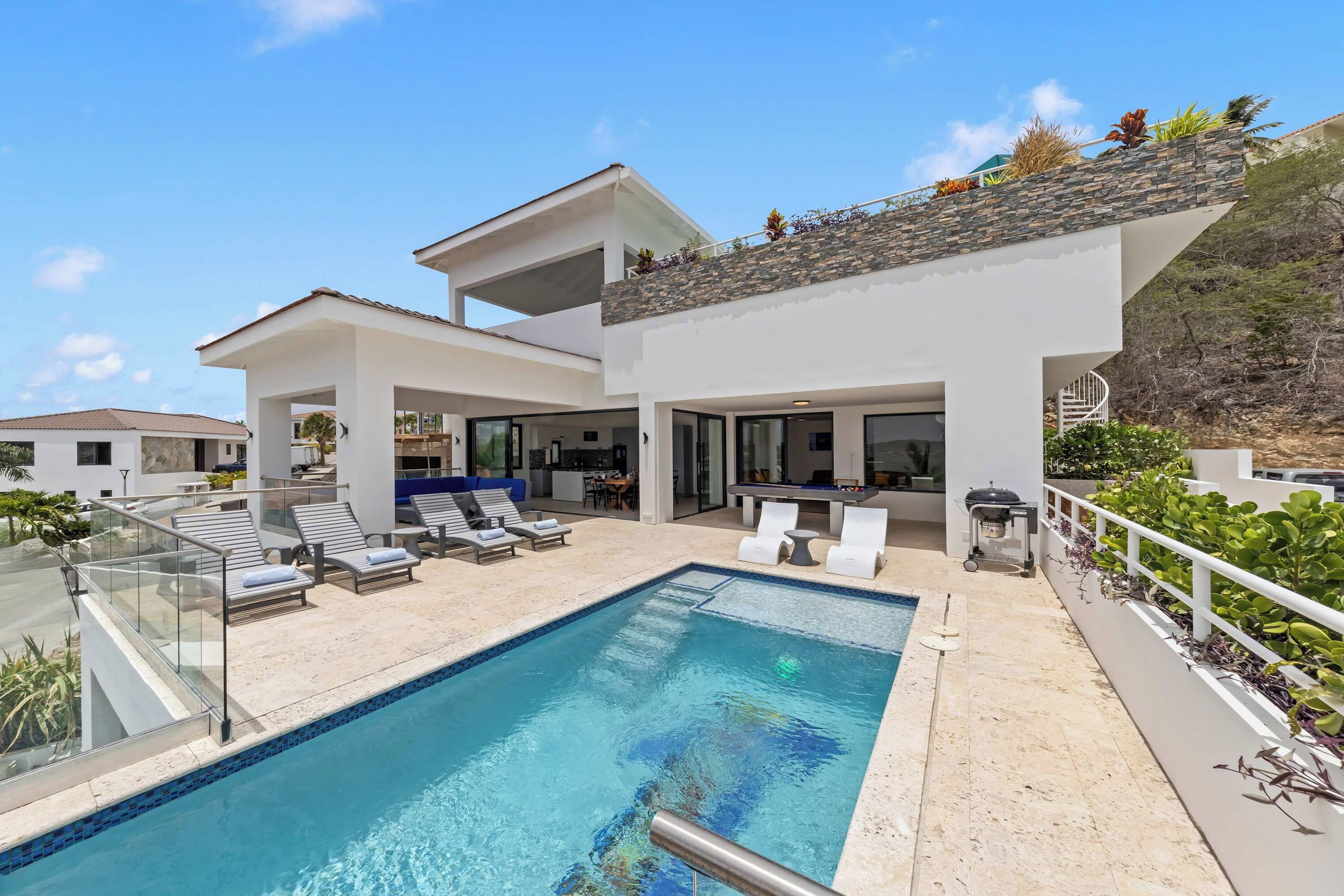 Ein modernes, zweistöckiges Haus mit Swimmingpool, Sitzgelegenheiten im Freien und Küchenbereich.