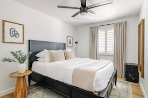 A bedroom with a bed, a ceiling fan, a potted plant, a bedside table, and a window with curtains.