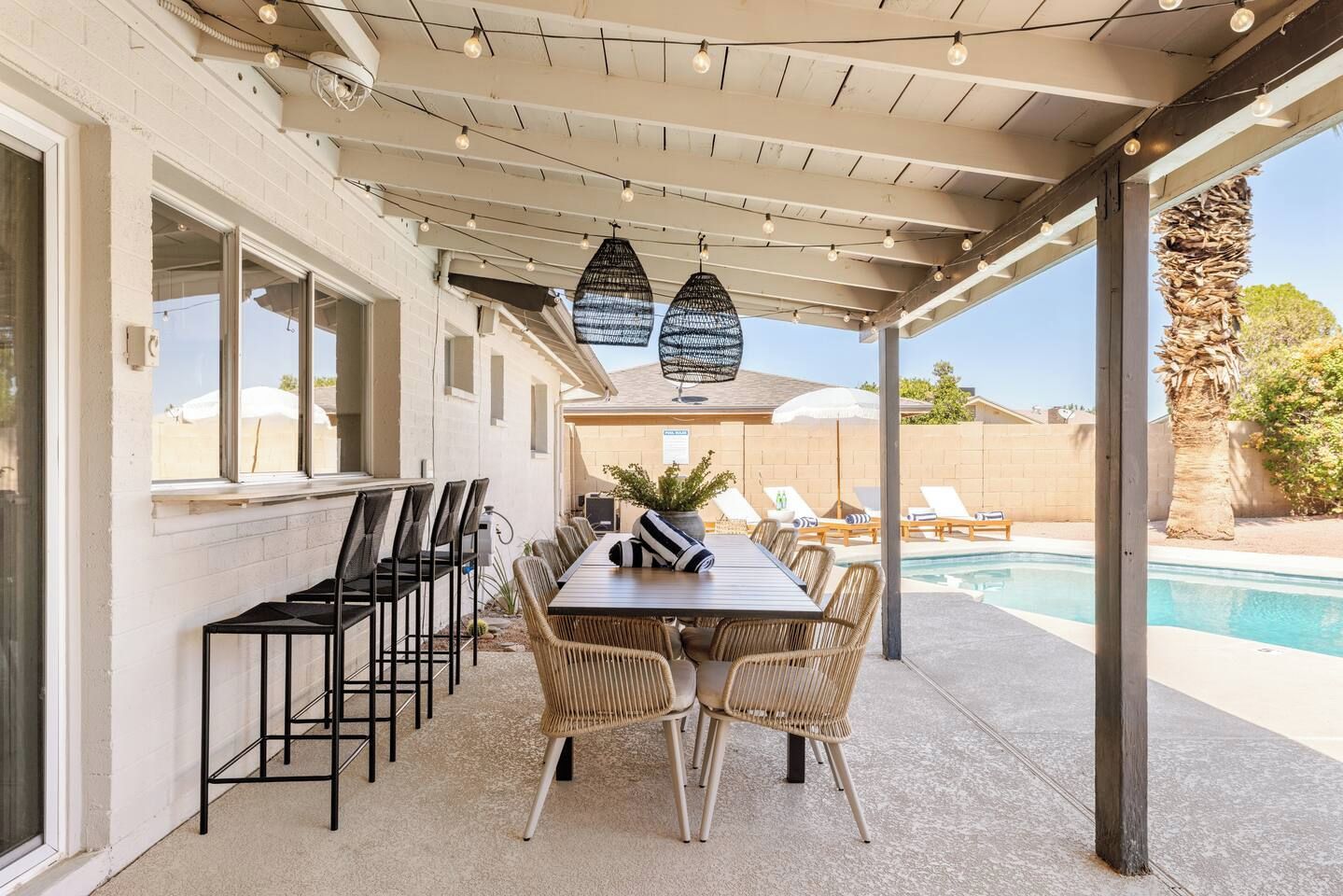 An outdoor patio with a pool, featuring a long dining table with chairs, a pergola with string lights, and a view of the pool area.