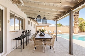 An outdoor patio with a pool, featuring a long dining table with chairs, a pergola with string lights, and a view of the pool area.