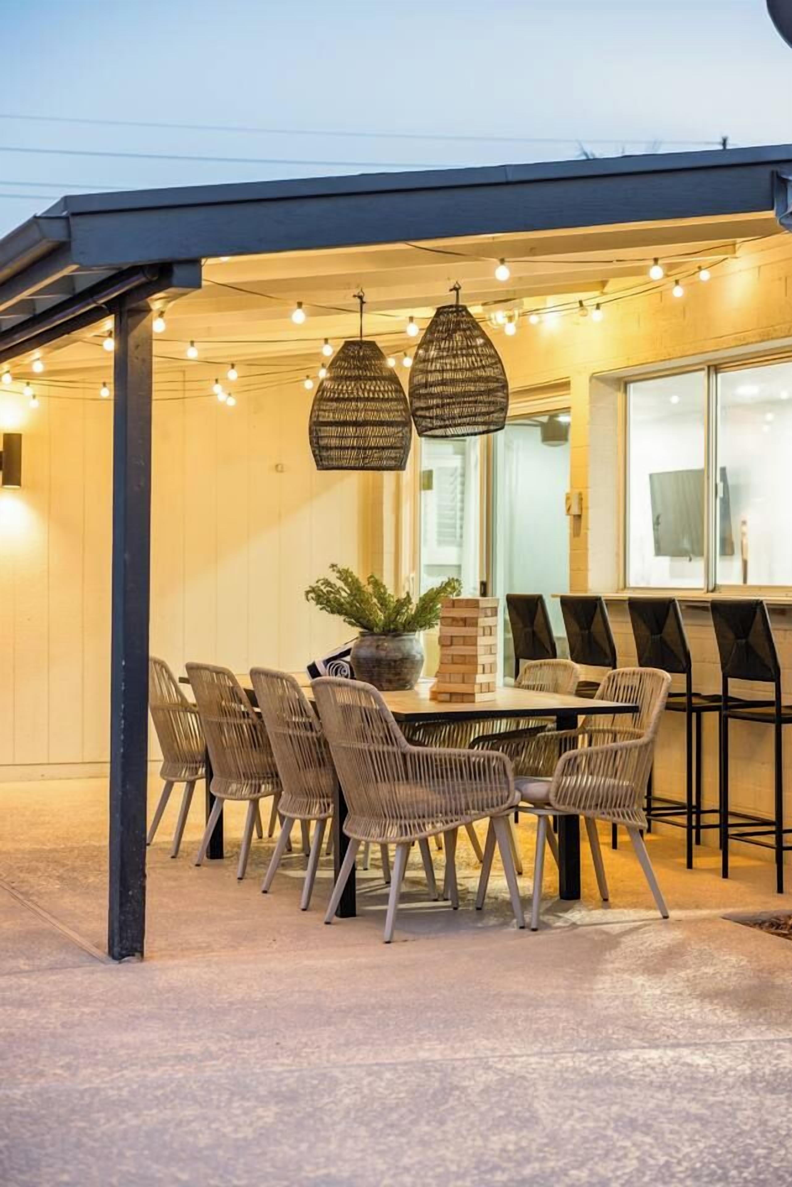 An outdoor dining area with wicker chairs and a table, under a covered structure with string lights.