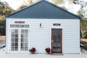 A small building with a metal roof, two doors labeled 'Reading Room' and 'Laundry,' and two potted plants with red flowers. - Djindarup Retreat Lakeview Tiny House 4 (Margaret River)