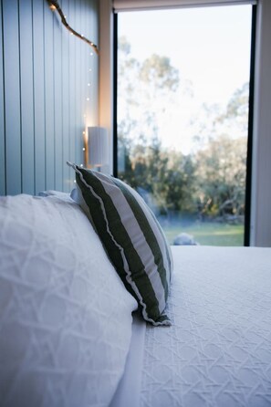 A bed with white bedding and a green striped pillow, a bedside lamp, and a view of trees through a window. - Djindarup Retreat Lakeview Tiny House 4 (Margaret River)