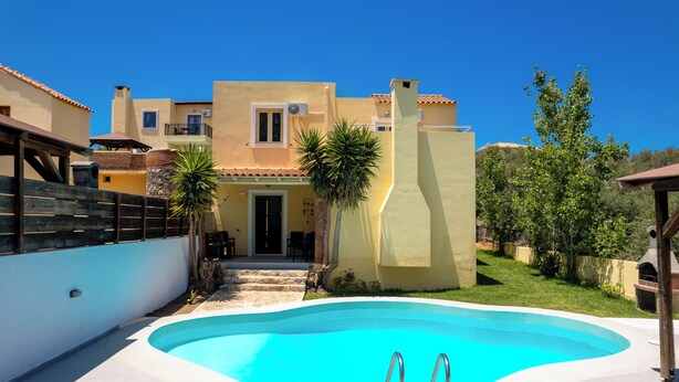 A swimming pool with lounge chairs, a building with a balcony, and a clear blue sky. - Villa Thalia (Χερσόνησος)