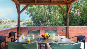 An outdoor patio with a table set for two, surrounded by brick walls and wooden beams, overlooking a lush green landscape. - Villa Thalia (Χερσόνησος)