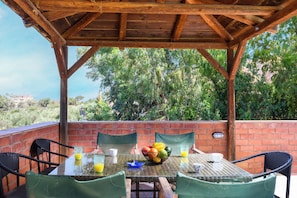 An outdoor patio with a table set for two, surrounded by brick walls and wooden beams, overlooking a lush green landscape. - Villa Thalia (Χερσόνησος)