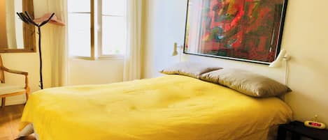 A bedroom with a bed, a chair, a window, and a painting on the wall.