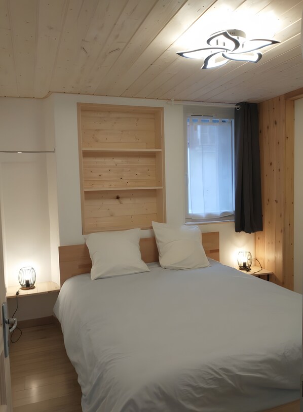 A bedroom with a wooden ceiling, a bed with white bedding, two bedside tables with lamps, and a window with a curtain.