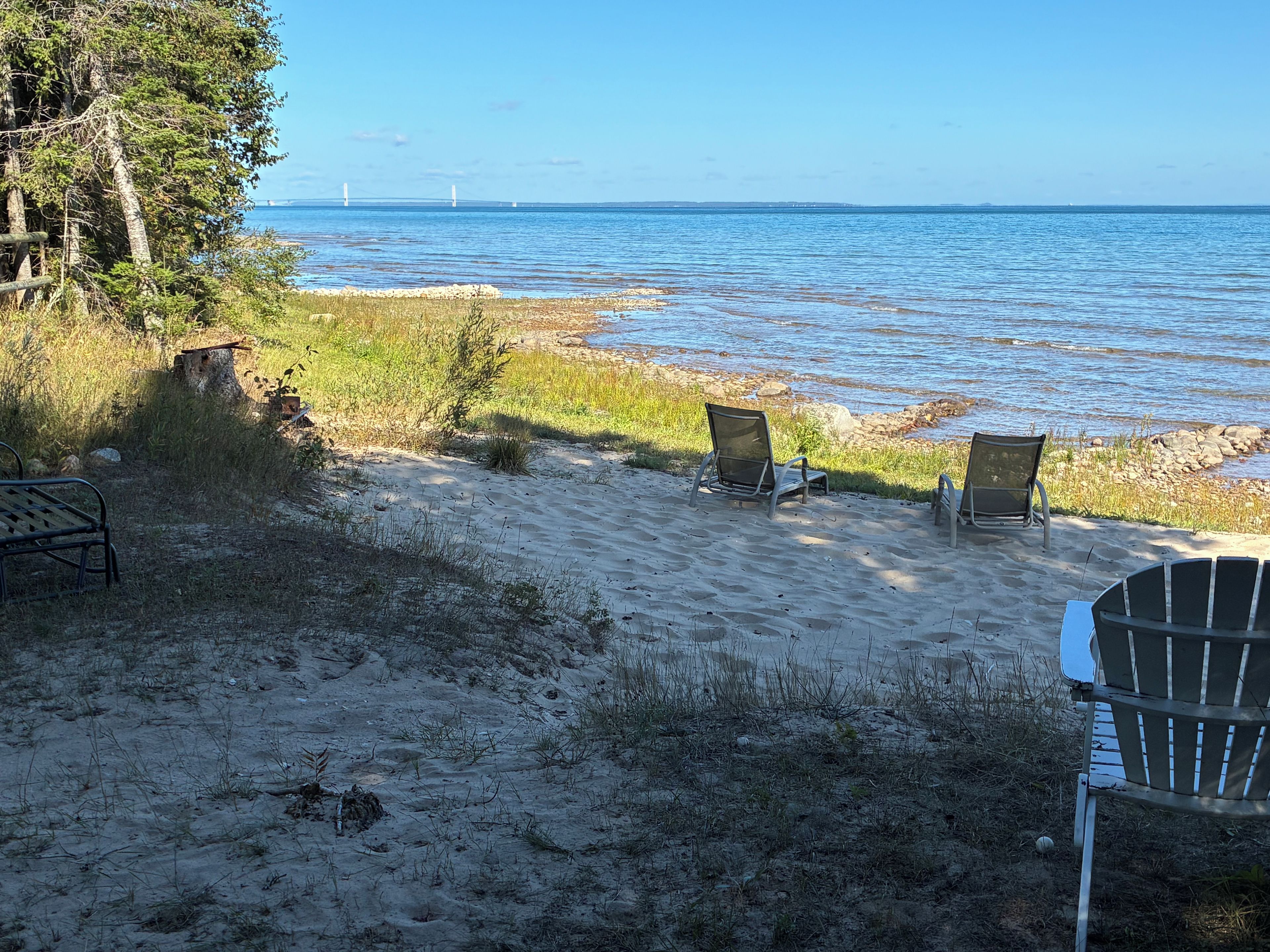 A beach with two chairs facing the water, a bench, and a tree line in the background.